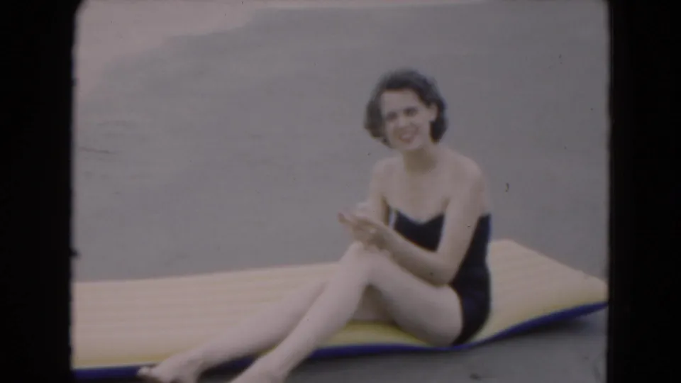 Closer 8mm frame of a smiling woman in a black one-piece swimsuit seated on a yellow-and-blue raft on a Florida beach.