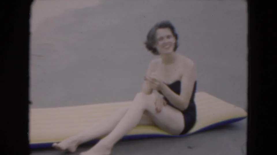 A smiling woman in a dark one-piece swimsuit sits sideways on a yellow-and-blue raft beside the ocean in supported 1975 Florida footage.
