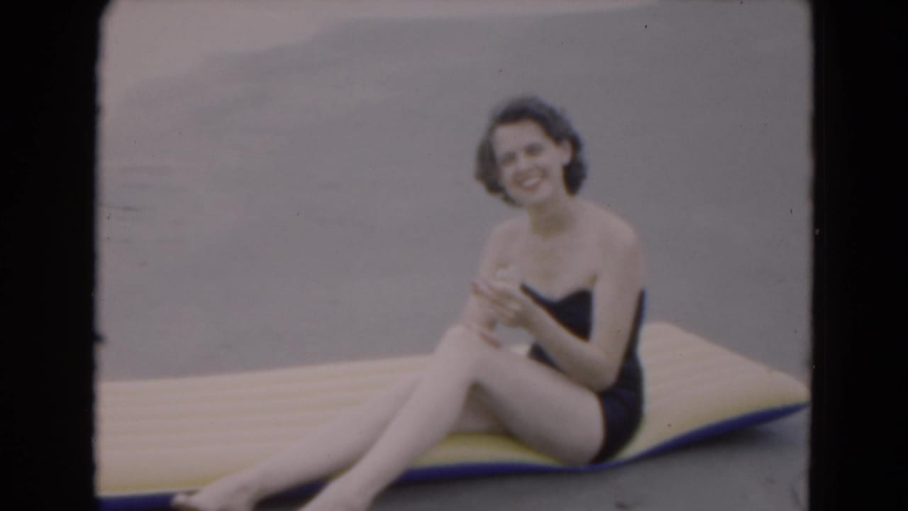 1975 Florida 8mm frame of a woman in a dark one-piece swimsuit smiling while seated on a yellow and blue inflatable board by the beach.