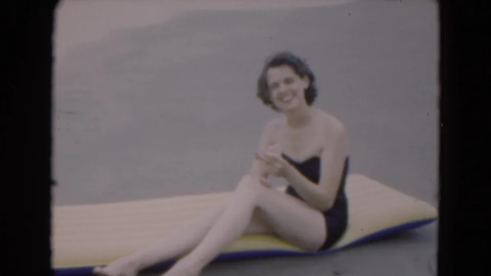 1975 Florida 8mm frame of a woman in a dark one-piece swimsuit smiling while seated on a yellow and blue inflatable board by the beach.