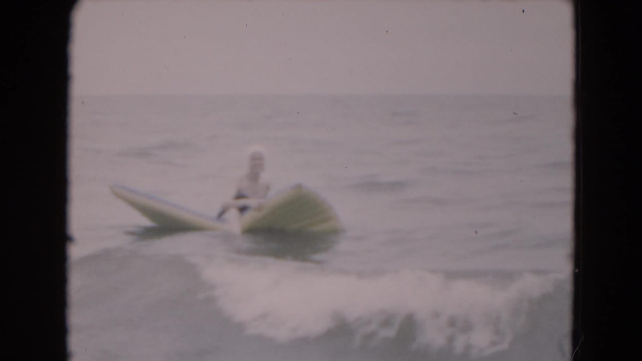 1975 Florida beach footage frame showing a woman in a white swim cap and dark swimsuit floating on a yellow board in gentle surf.