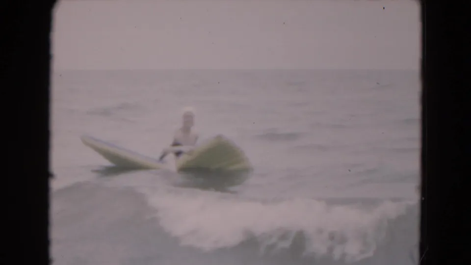 1975 Florida beach footage frame showing a woman in a white swim cap and dark swimsuit floating on a yellow board in gentle surf.