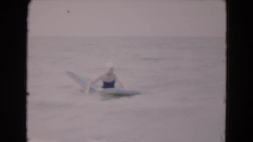 Soft 8mm frame from 1975 Florida showing a woman in a swim cap beside a yellow board, partly obscured by shallow ocean water.