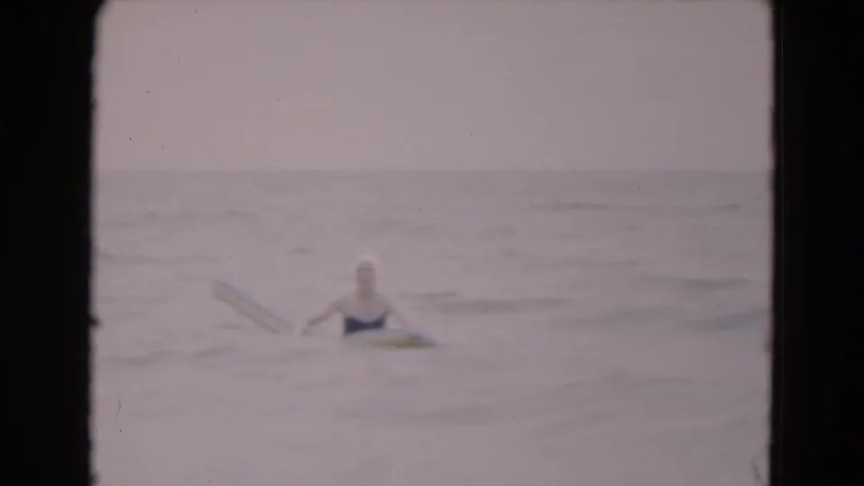 1975 Florida 8mm frame of a woman in a swim cap floating farther out on a yellow board amid low, calm ocean waves.