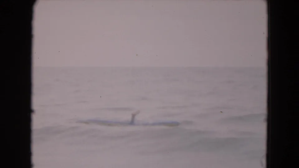 1975 Florida beach footage frame with a raft drifting through gentle water and a distant person partly visible.