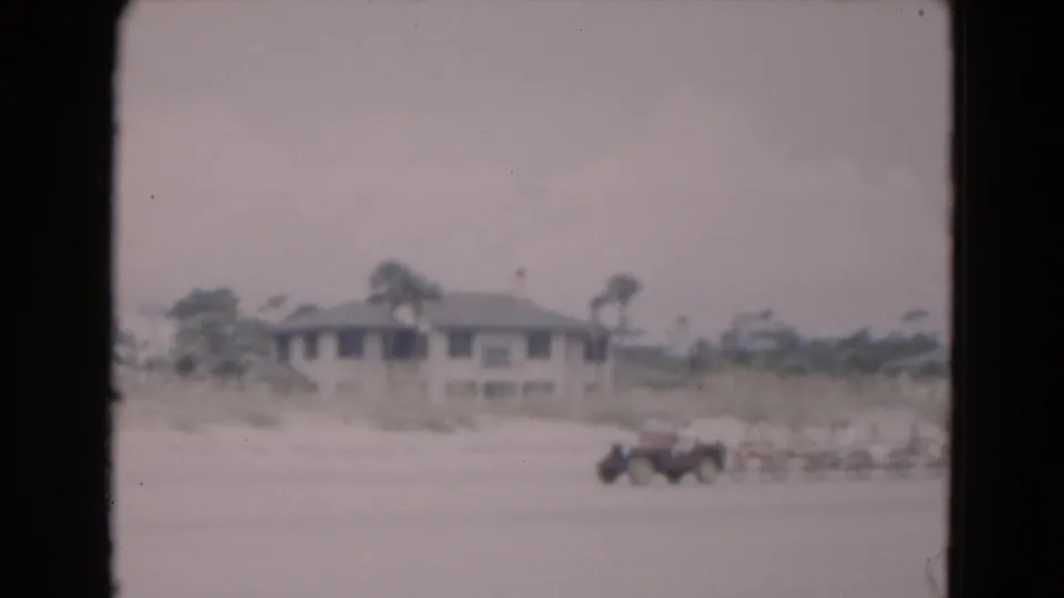 1975 Florida 8mm frame showing a low beach house, palms, dunes, and a car parked on the sand.