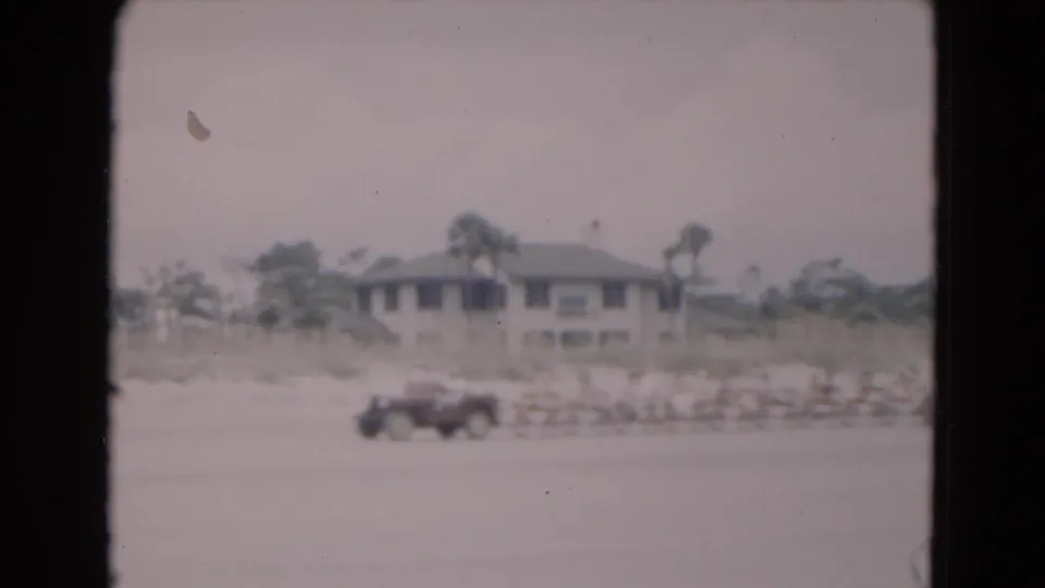 1975 Florida home-movie frame with a car on the sand and a beach house set back behind dunes and palms.