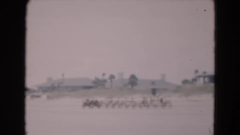 1975 Florida 8mm frame showing a distant row of beachgoers on a sandy beach, with low coastal buildings and palm trees in the background.