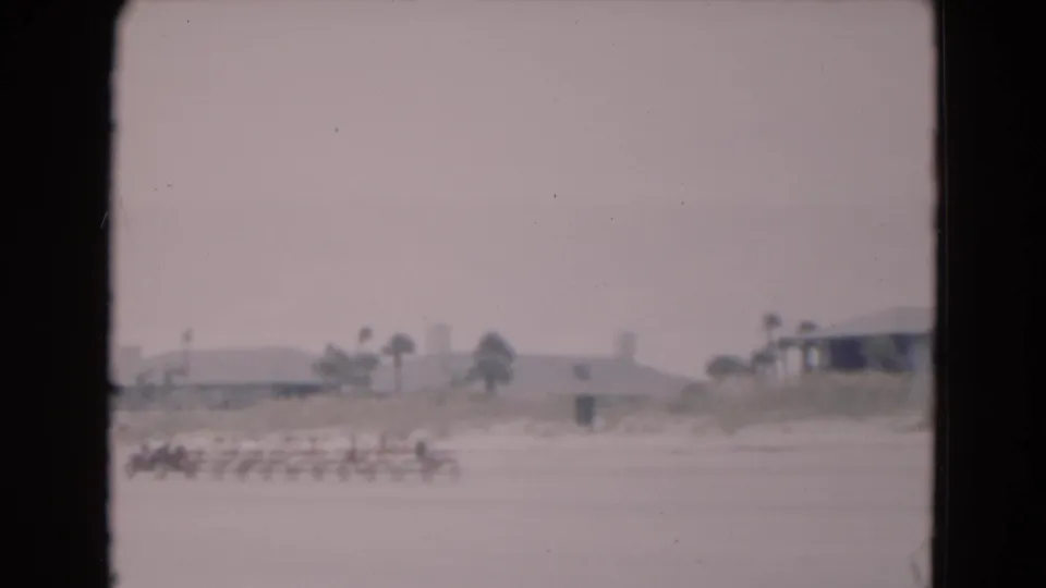 Distant 1975 Florida beach view with seated people arranged in a row on sand and low buildings with palms behind them.