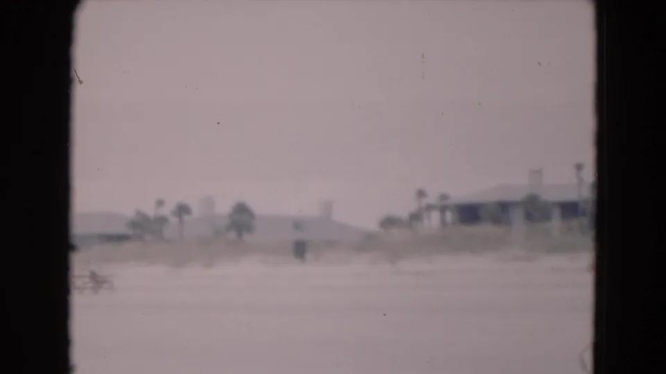 Soft 8mm beach frame from Florida in 1975, showing open sand, distant palms, low buildings, and small figures near the side of the view.