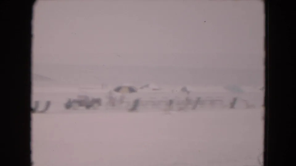 Soft 1975 8mm frame showing distant base-like structures across a pale icy landscape, with dark film borders and haze.