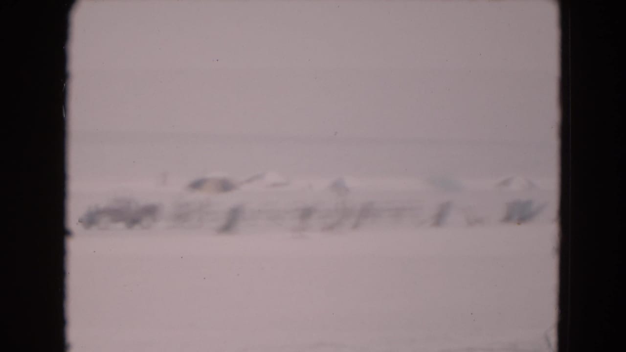1975 8mm frame with a hazy view of distant structures and possible vehicles or equipment on a flat icy field.
