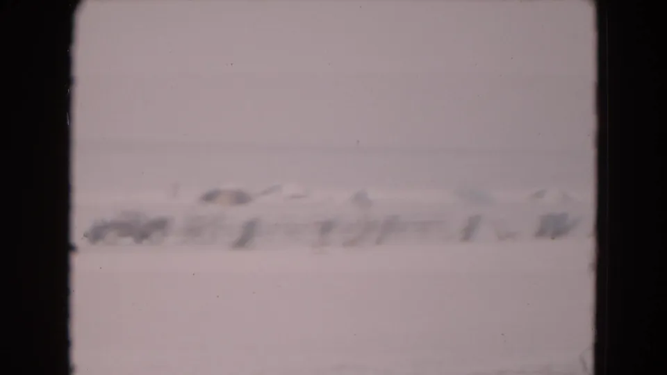 1975 8mm frame with a hazy view of distant structures and possible vehicles or equipment on a flat icy field.