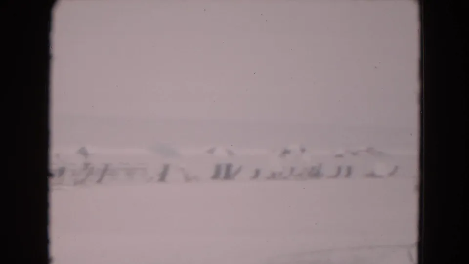 Soft 1975 8mm frame showing faint low buildings or equipment across a snow-covered horizon, with heavy film borders.