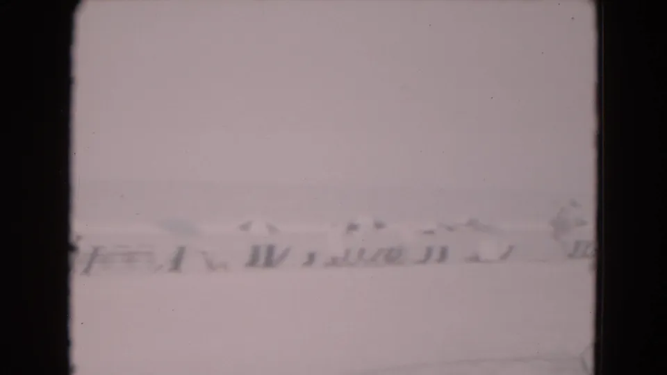 1975 8mm frame with a blurred line of distant structures or equipment at the edge of a pale icy landscape.