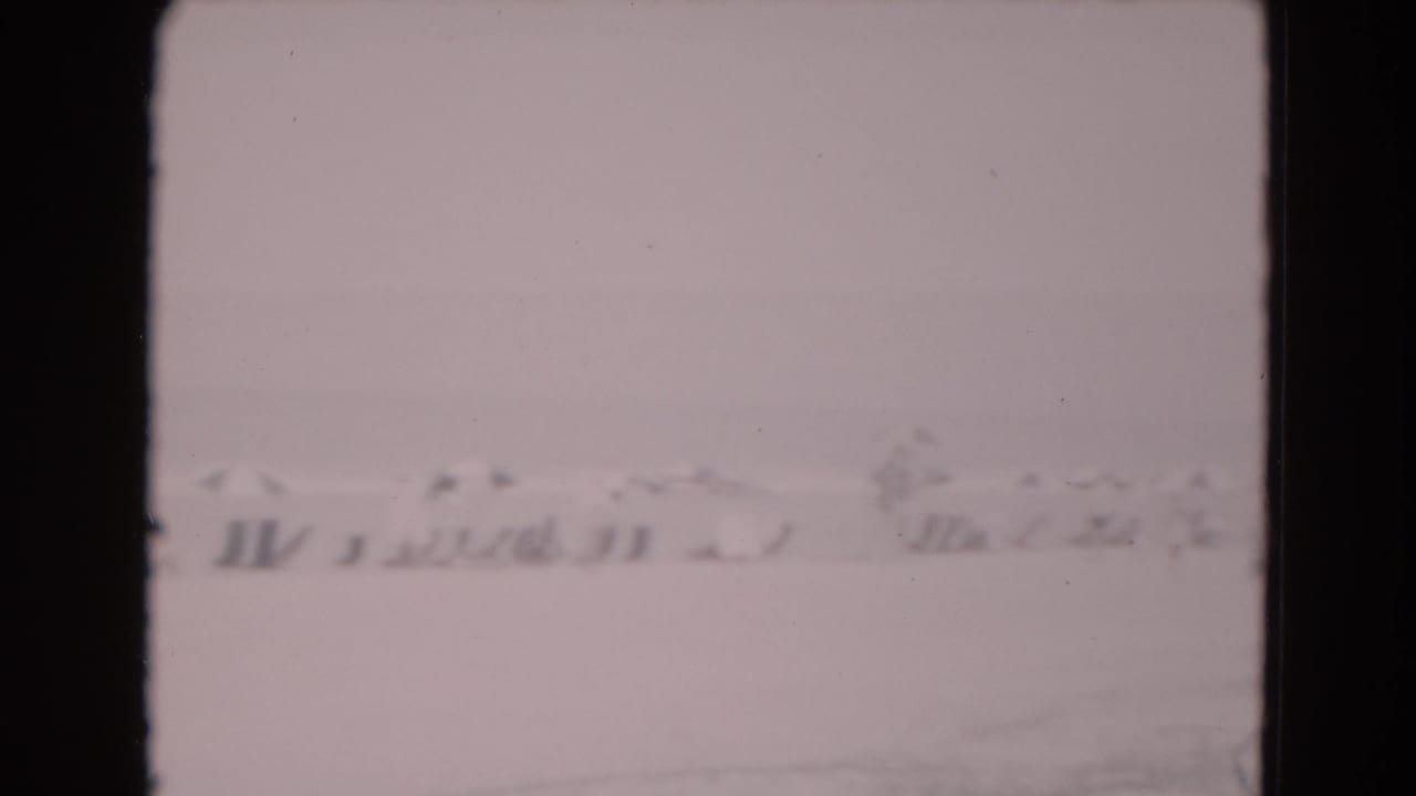 Pale snow-covered or frost-like landscape with low indistinct structures under a gray sky.