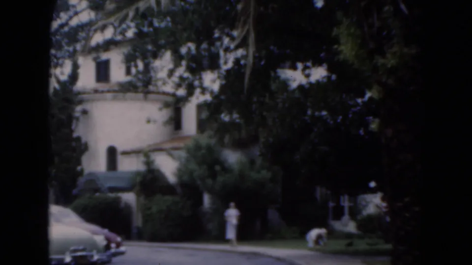 1975 Florida 8mm frame showing a distant person in light clothing near a light-colored residence with palms, shrubs, and lawn.