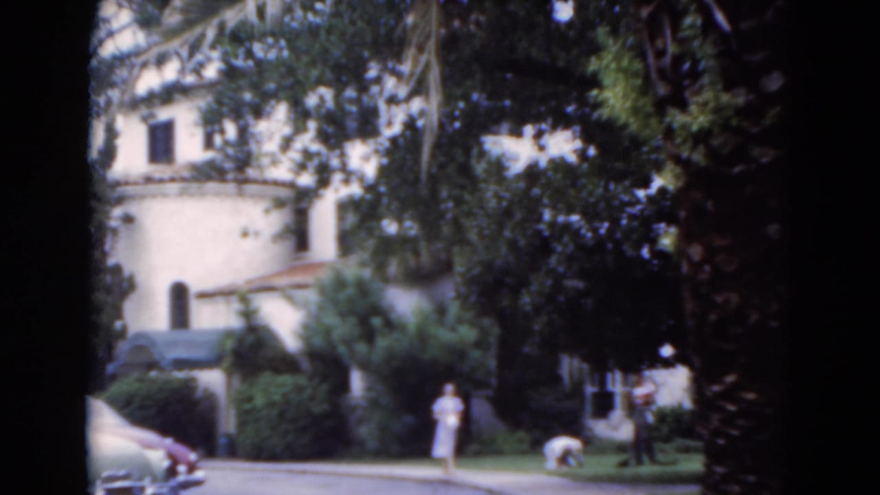 1975 Florida home-movie frame with a woman on the walkway, another person bending near the lawn, and a stucco building behind trees.