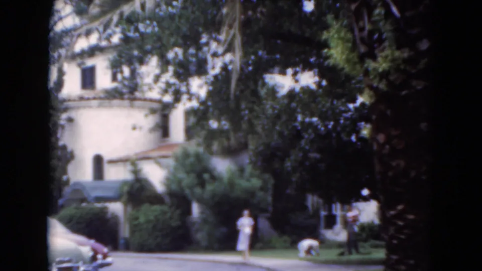 1975 Florida home-movie frame with a woman on the walkway, another person bending near the lawn, and a stucco building behind trees.