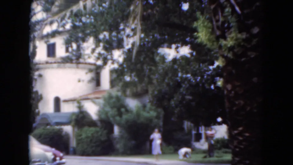 Soft 8mm 1975 Florida frame showing a distant woman near a walkway, a person by the lawn, and a pale stucco building partly hidden by trees.
