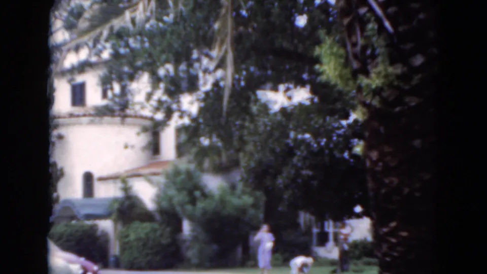 1975 Florida 8mm frame with a woman in a light dress near the walkway, another person on the grass, and a rounded stucco building in the background.