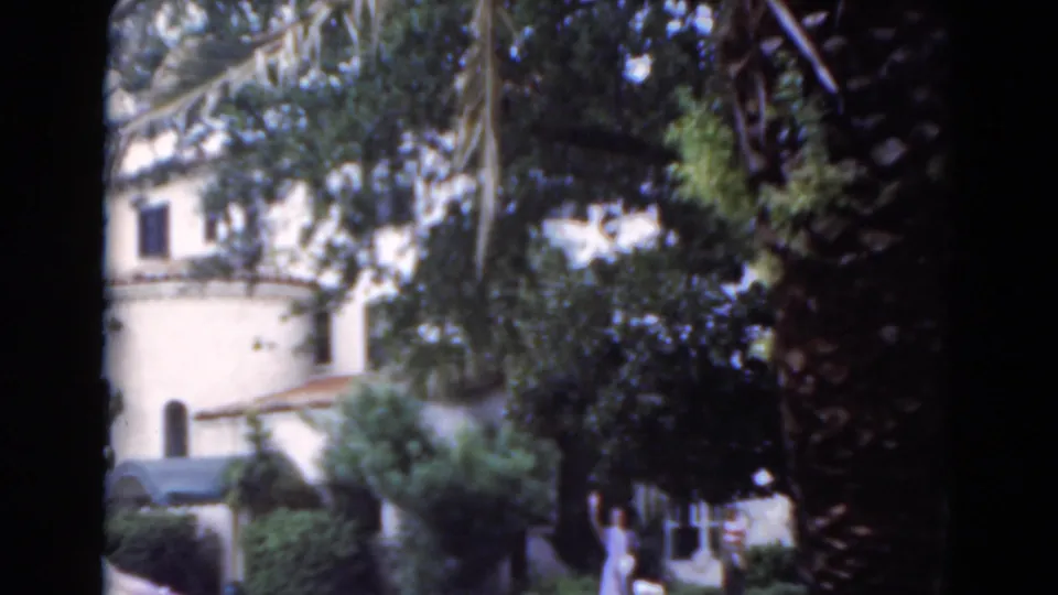Soft 8mm view of a Florida residential garden in 1975, with stucco walls, palms, shrubs, and a distant person near the lawn.