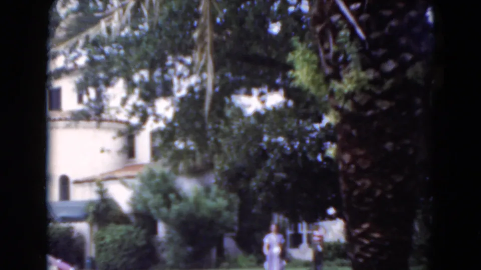 1975 Florida home-movie frame of a woman in light clothing and a small dog on a lawn below a white stucco house with palms.