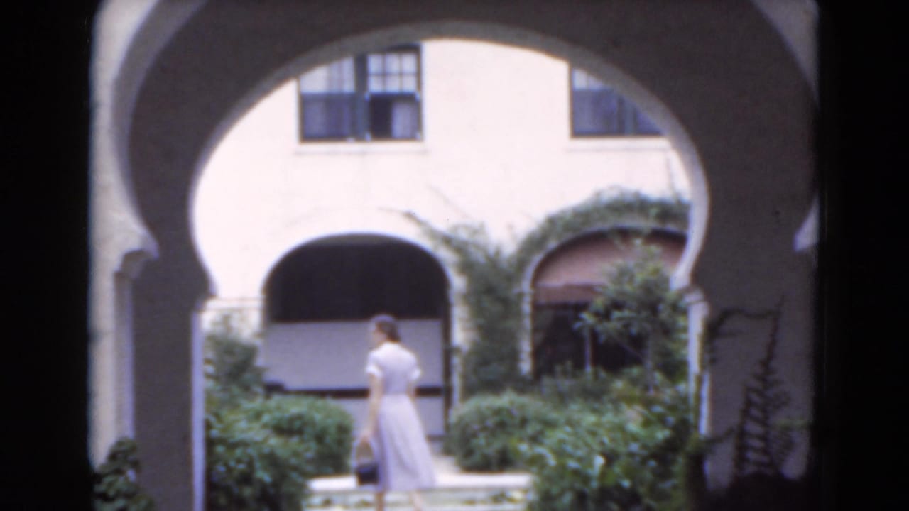 Woman in a pale dress walks beneath arched courtyard openings beside shrubs.