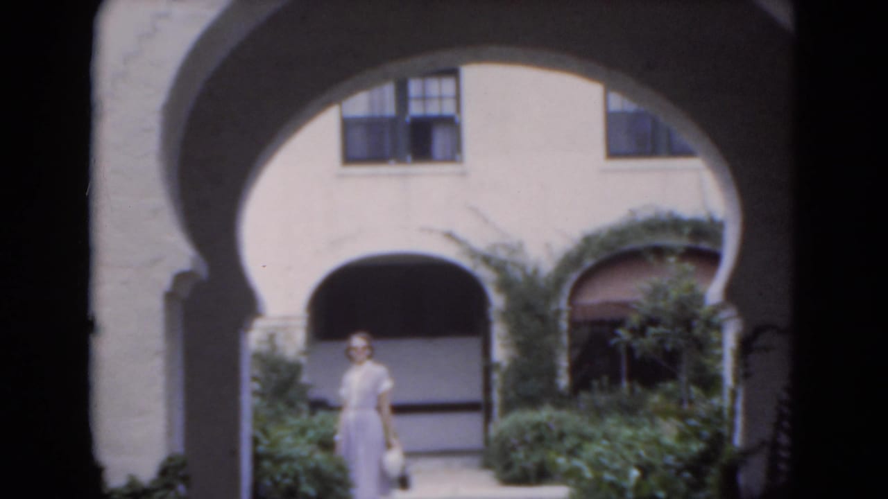 Woman stands near the center of an arched courtyard with greenery around her.