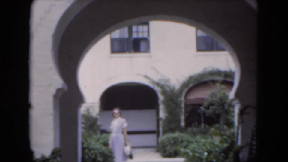 The woman faces the camera in a Florida courtyard framed by stucco arches, shrubs, and dark upper windows.
