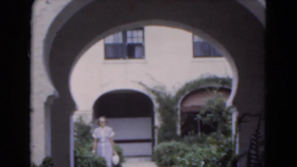 A woman in a light dress stands partly in shade under a broad arch in the 1975 Florida courtyard scene.