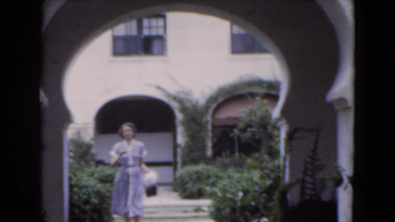 A woman in a light dress walks toward the camera through a Florida courtyard framed by white stucco arches and dense planting.