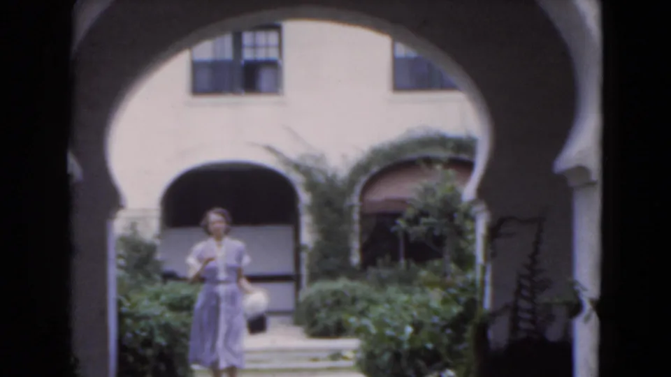 A woman in a light dress walks toward the camera through a Florida courtyard framed by white stucco arches and dense planting.