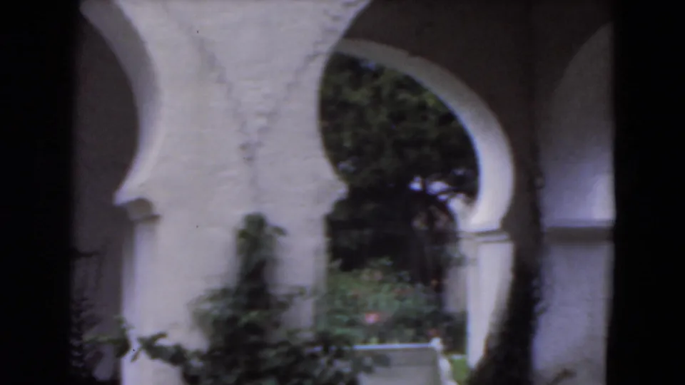 The handheld 8mm view shifts across a white arched arcade and garden planting in the Florida courtyard setting.