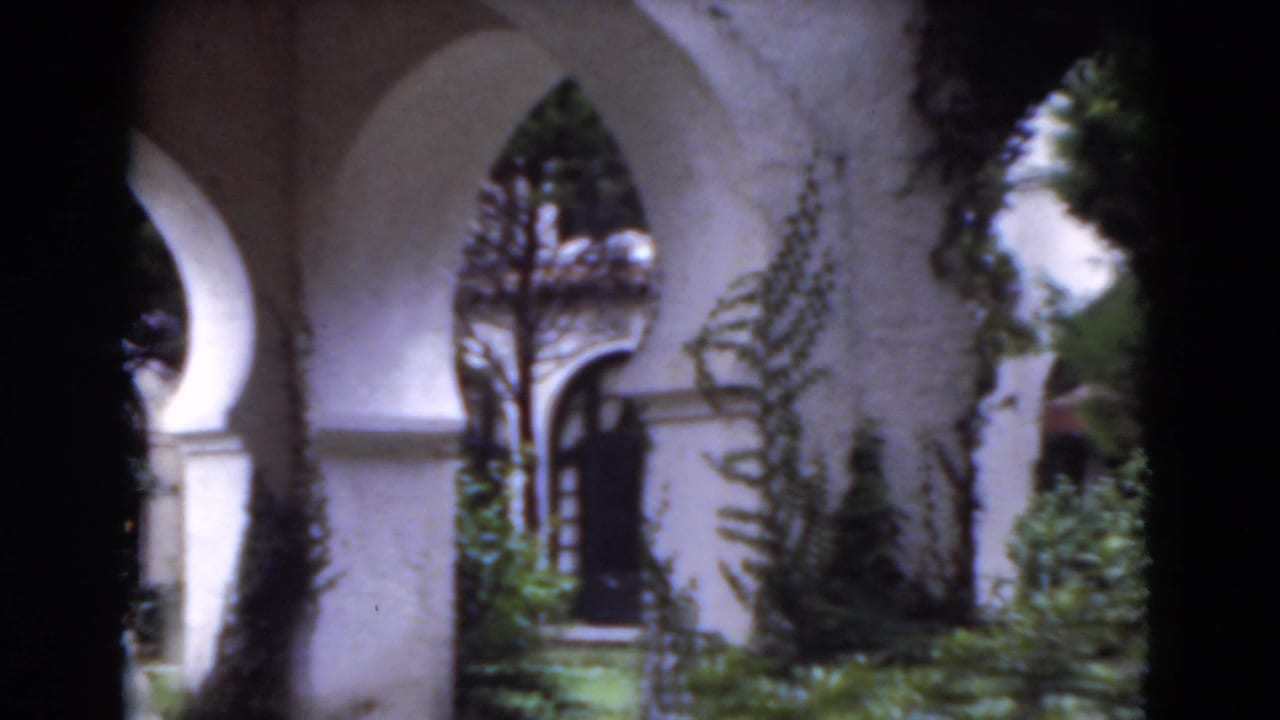 White stucco arcade with arches, vines, shrubs, and lawn in a garden setting.