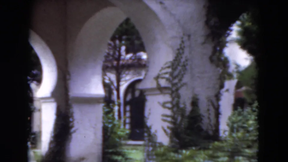 1975 Florida 8mm frame showing a white stucco arched walkway, climbing plants, shrubs, and green lawn.