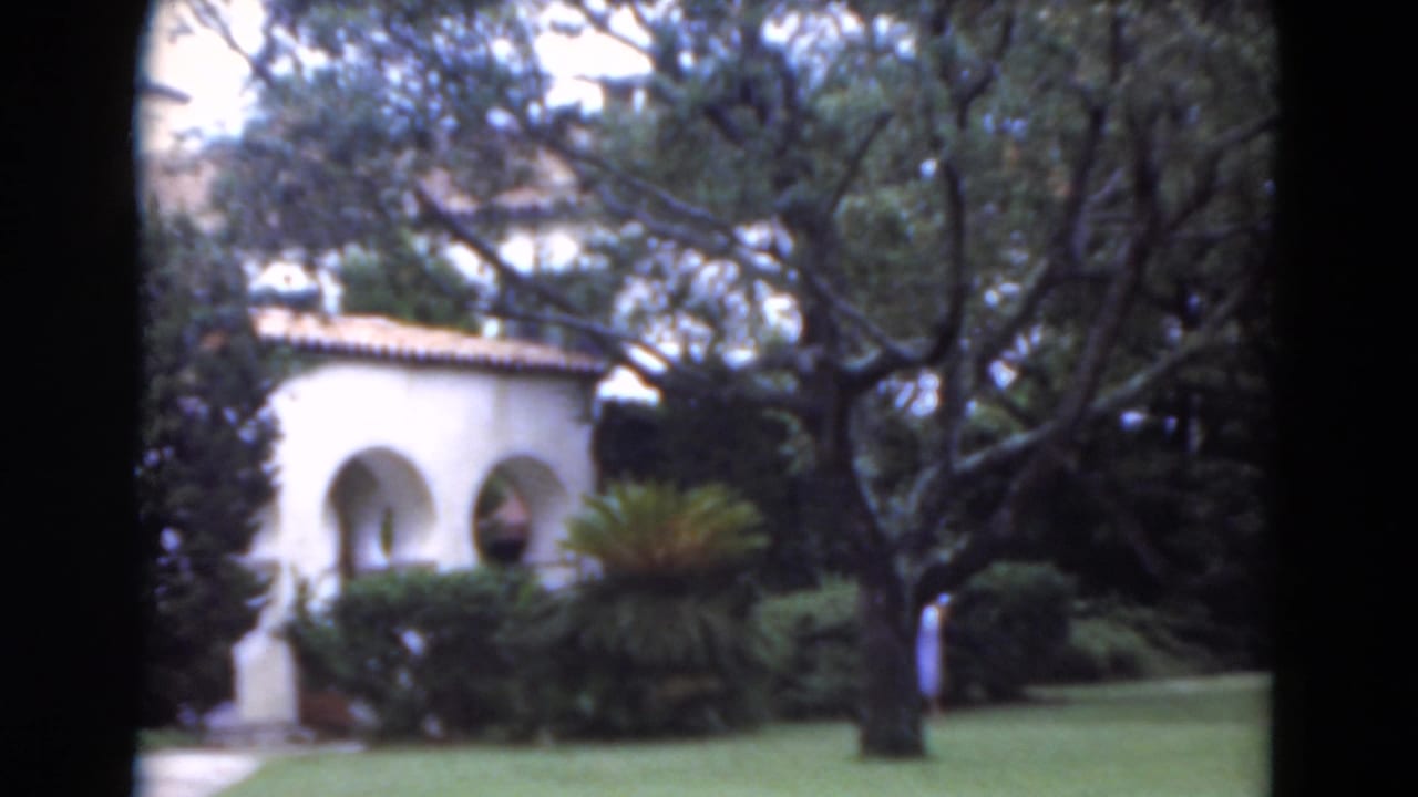 Garden lawn with white stucco arches, trees, shrubs, and a distant woman near the right edge.