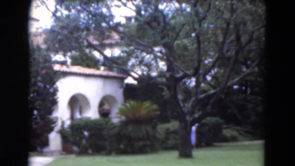 1975 Florida garden view with a white stucco building, arched openings, mature trees, shrubs, and a distant woman on the lawn.