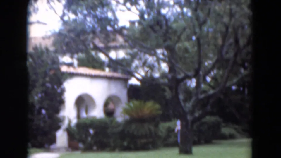 Soft 8mm frame of a landscaped Florida lawn, white arched building, trees, shrubs, and a partly visible woman at the edge.