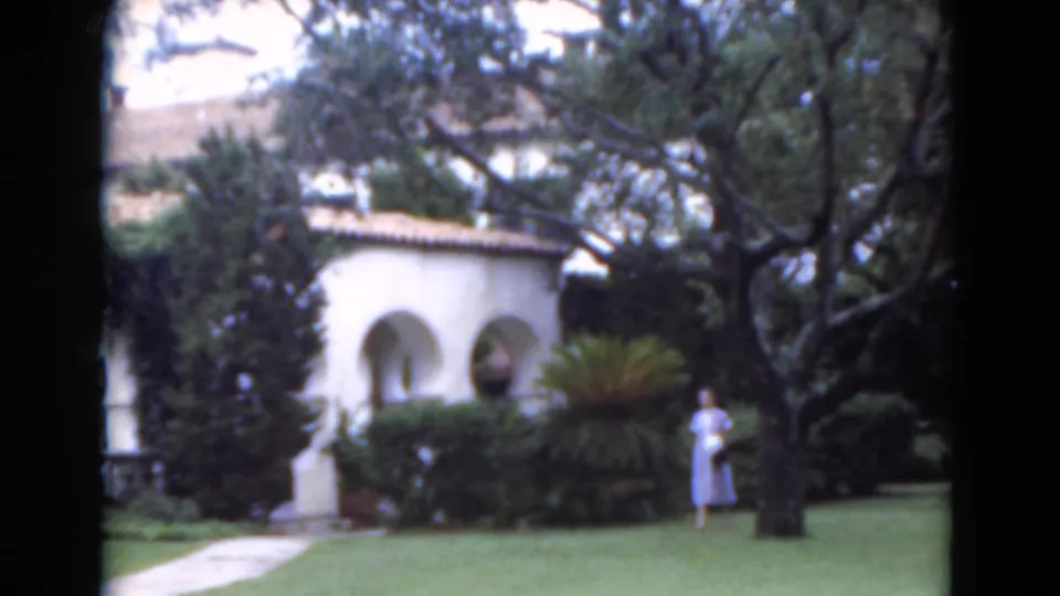 1975 Florida 8mm frame with a woman in a light dress on the lawn beside trees, shrubs, a path, and white stucco arches.