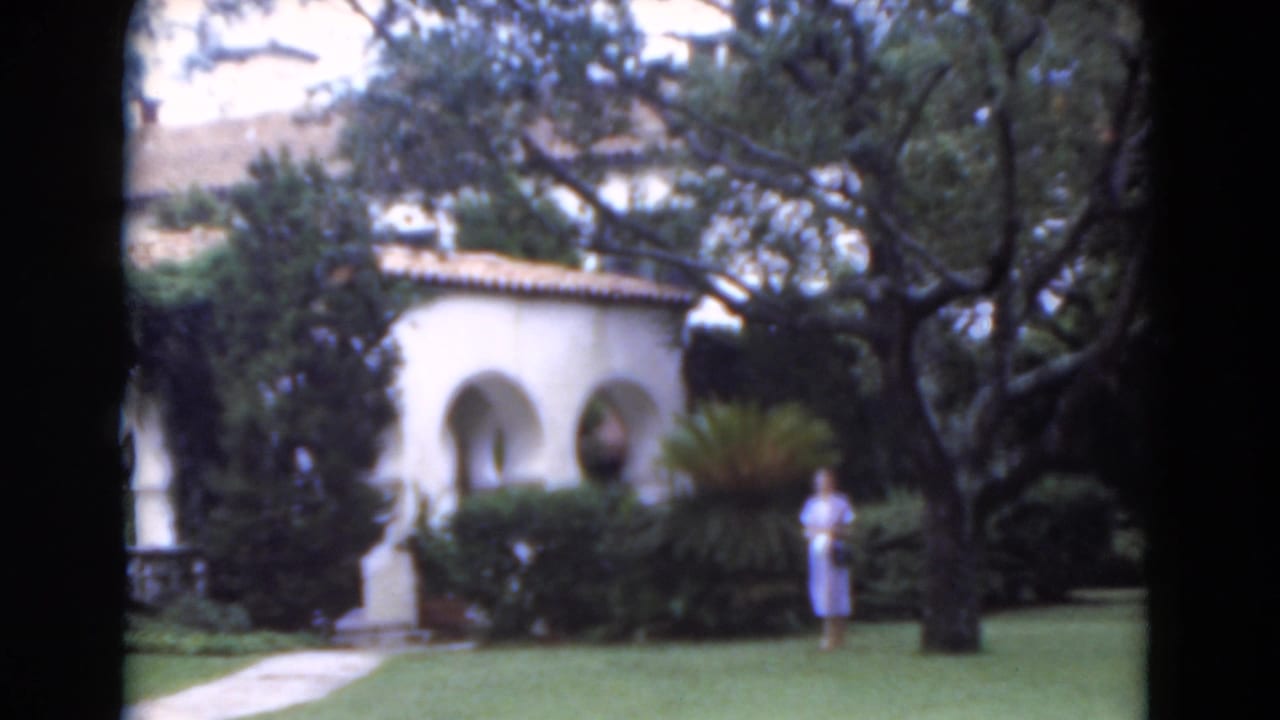 Woman in a light blue dress standing on a lawn near white stucco arches and trees.
