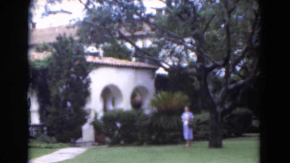 1975 Florida 8mm frame showing a woman in a light blue dress on a manicured lawn beside white stucco arches and dense greenery.