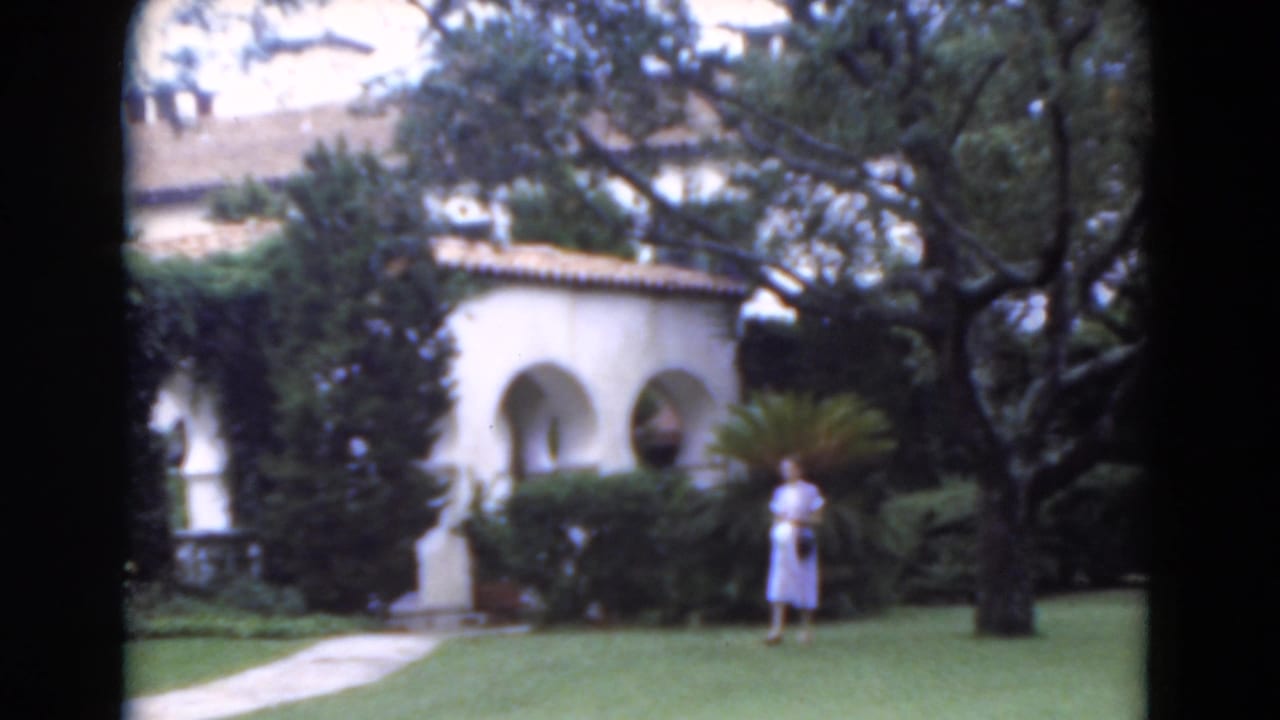 1975 Florida home-movie frame with a woman on a green lawn, framed by a white arched exterior, hedges, and mature trees.