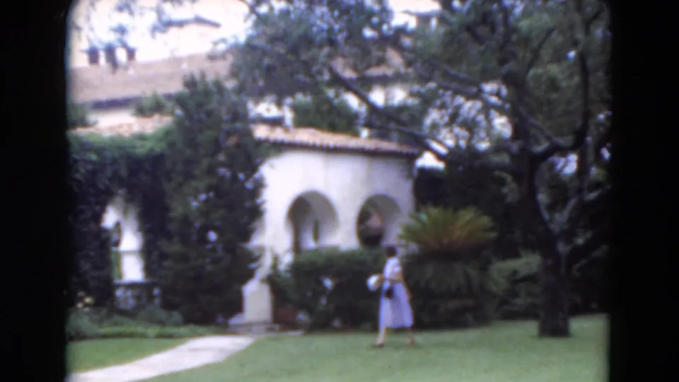 1975 Florida 8mm frame showing the woman walking left across the lawn, with stucco arches, shrubs, and trees in the background.