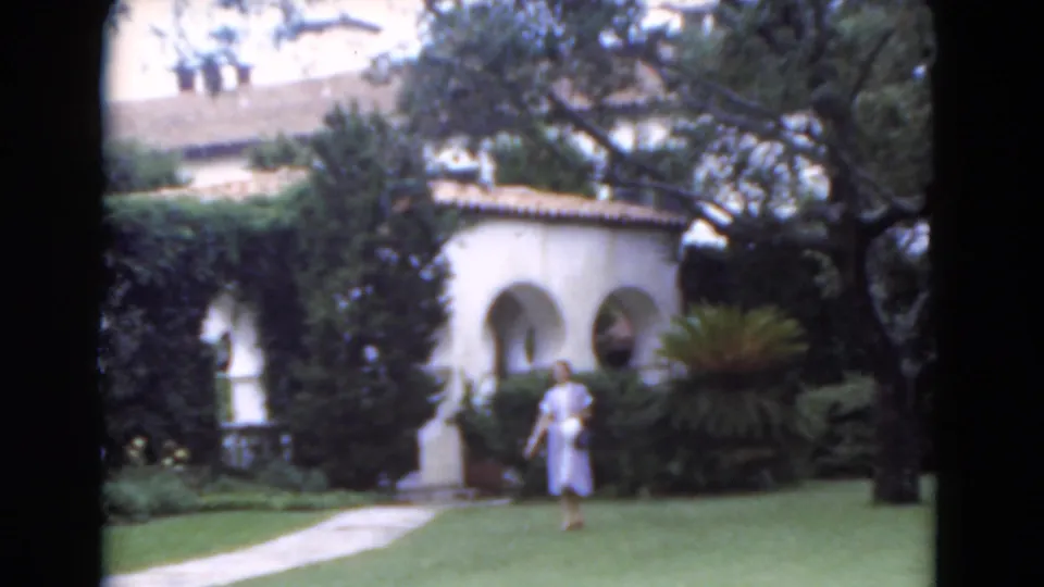 1975 Florida home-movie frame of a woman in a light blue dress crossing a manicured lawn near shrubs, trees, and white arches.