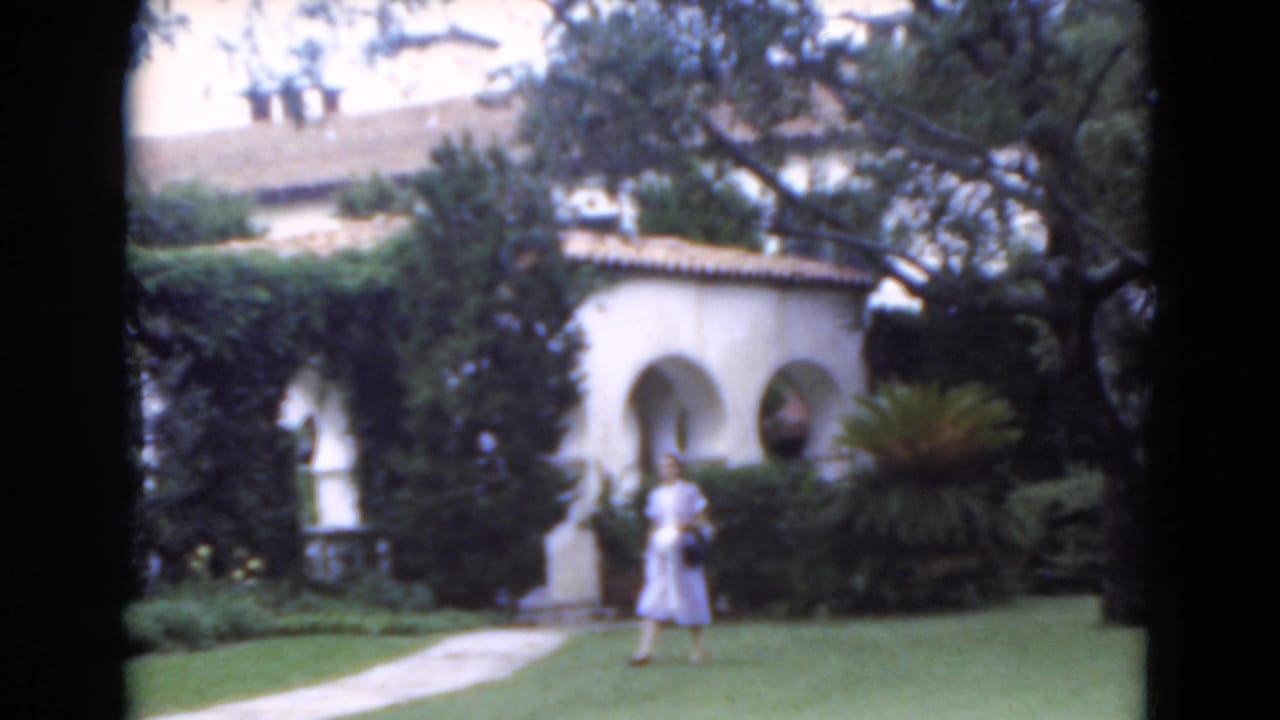 1975 Florida 8mm frame of a woman crossing a manicured lawn toward a paved walkway beside stucco arches and dense garden plantings.