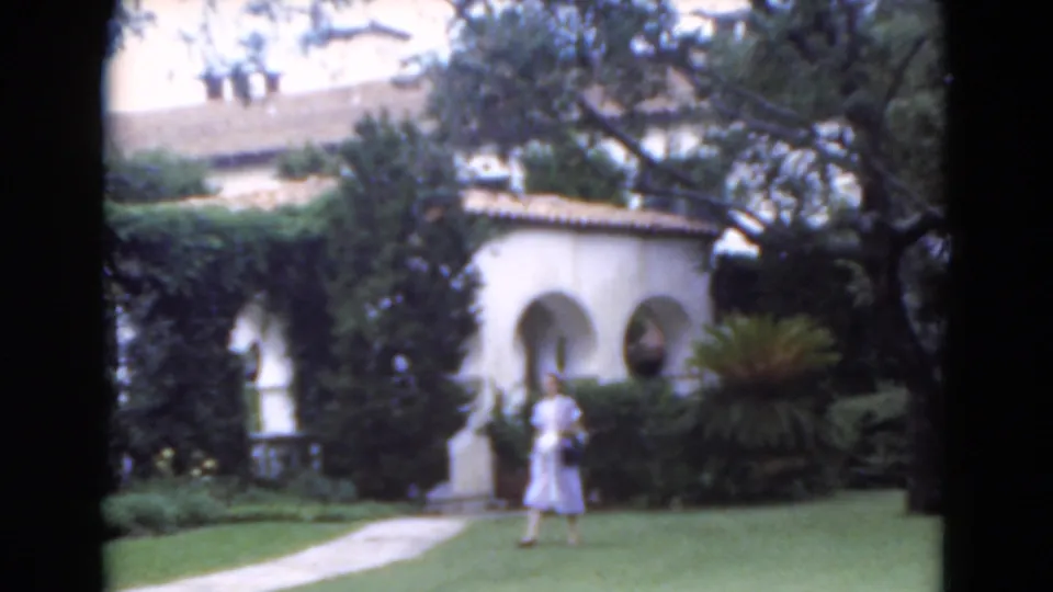 1975 Florida 8mm frame of a woman crossing a manicured lawn toward a paved walkway beside stucco arches and dense garden plantings.