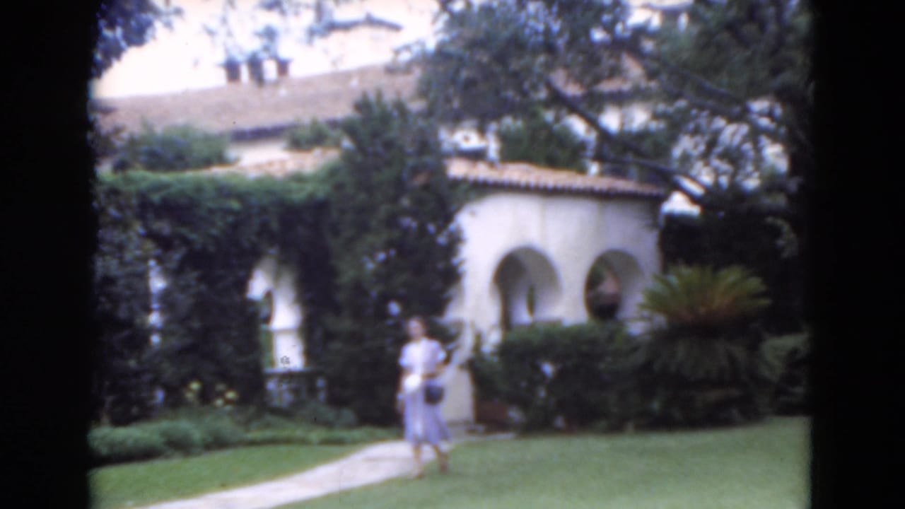1975 Florida home-movie frame showing a woman near a paved garden path in front of Spanish-style arches and dense landscaping.