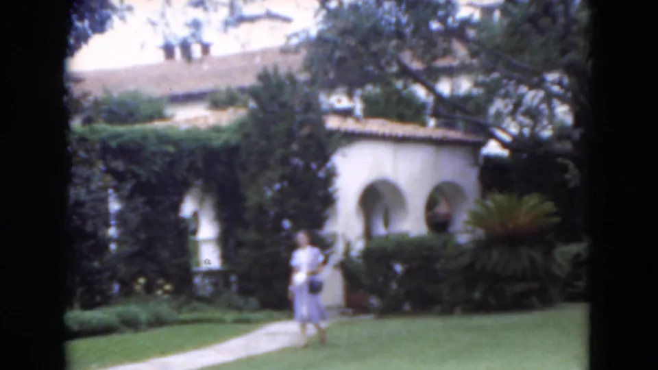 1975 Florida home-movie frame showing a woman near a paved garden path in front of Spanish-style arches and dense landscaping.