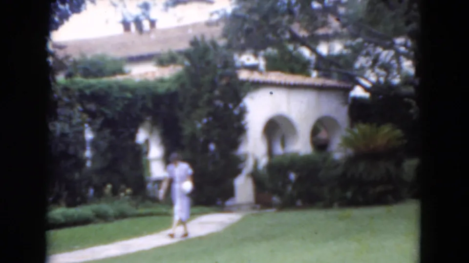 1975 Florida 8mm frame of a woman moving along a curved walkway past a manicured lawn and arched stucco garden wall.